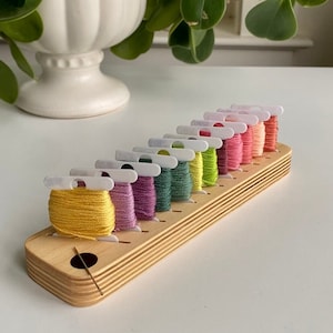 May include: A wooden embroidery floss organiser with 12 slots. The slots are filled with colourful embroidery floss in shades of yellow, green, purple, pink and red.