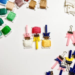 May include: A collection of colorful embroidery floss and accessories, including pink, yellow, and blue binder clips. Various spools of thread in shades of green, brown, and pink are arranged on a white surface, ideal for crafting and embroidery projects.