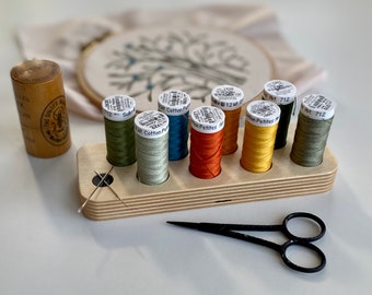 May include: A wooden embroidery floss organiser with eight spools of thread in various colours, including green, orange, yellow, and white. The organiser has a label that reads "Art. 712" and "Petites A".