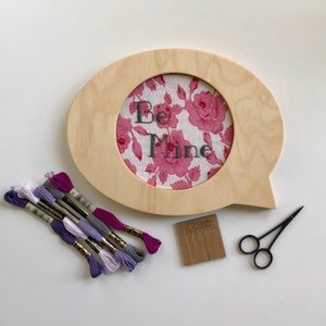 May include: A wooden speech bubble frame with a floral fabric insert that reads "Be Mine." Embroidery floss in shades of purple and gray, small scissors, and a Modern Koala tag are also present.