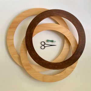 May include: Three wooden hoops, one brown and two natural wood, are stacked on top of each other. A pair of scissors and a spool of green yarn are in the centre of the hoops.