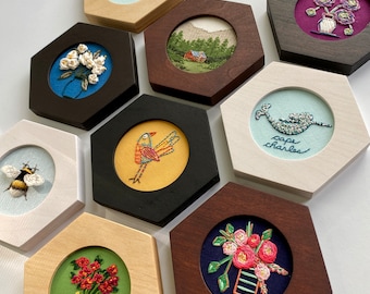 May include: A collection of 10 octagonal wooden frames with embroidered designs. The designs include a bird, a bouquet of flowers, a mermaid, a house, and a floral arrangement. The frames are all different colors, including black, brown, white, and natural wood.
