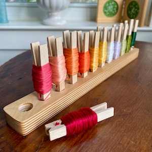 May include: A wooden embroidery floss organizer with 12 slots, each holding a different color of floss. The floss is secured with wooden clothespins. A red floss and clothespin are lying on the table next to the organizer.