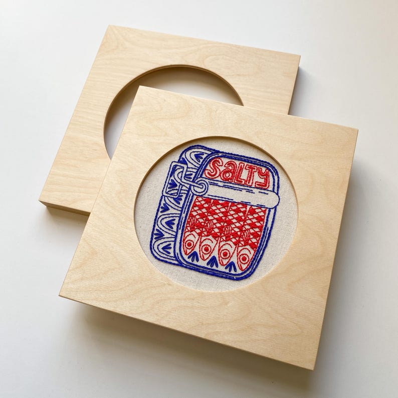 Puede incluir: Un marco de madera con una abertura circular, que muestra un dise&ntilde;o bordado en azul y rojo de una lata de sardinas con la palabra "SALTY" en ella.