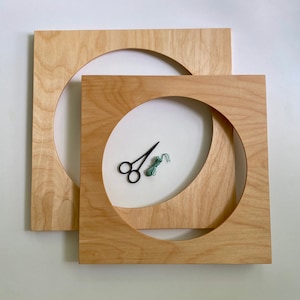 May include: Two square wooden embroidery hoops with a circular cutout, one larger than the other. Inside the smaller hoop, there are black scissors and a small bundle of green thread. The wood has a light, natural finish.