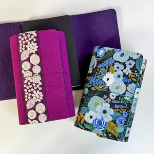 May include: Two journal covers. One is a deep magenta with a white floral embroidered design. The other is black with a floral pattern in shades of blue, white, and yellow. Both are closed with an elastic band.