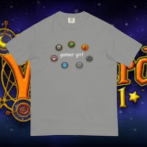 Może przedstawiać: Szary T-shirt z nadrukiem dziewczyny grającej w gry. Nadruk zawiera różne kolorowe ikony, w tym czaszkę, liść, płomień, chmurę, płatek śniegu i trójkąt.