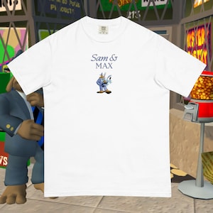 May include: White t-shirt featuring the text "Sam & Max" in a light blue font, with an illustration of the characters Sam and Max. Sam is wearing a suit and Max is holding a weapon. The shirt is made of a soft, comfortable material.
