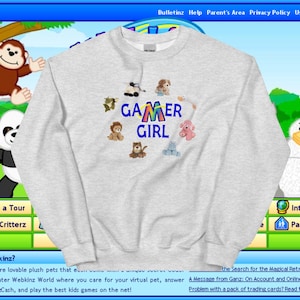 Peut inclure: Un sweat-shirt gris avec l'inscription "Gamer Girl" imprimée en lettres arc-en-ciel. Le sweat-shirt est décoré d'un cercle de peluches, dont un lion, un chat, une oie, un hippopotame, un chien et un singe.