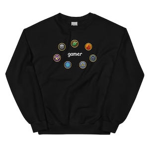 Peut inclure: Sweat-shirt noir avec le mot "gamer" en blanc, entouré de huit icônes circulaires colorées. Les icônes comprennent un crâne, une feuille, une flamme, un éclair, un flocon de neige, un triangle et deux autres symboles.