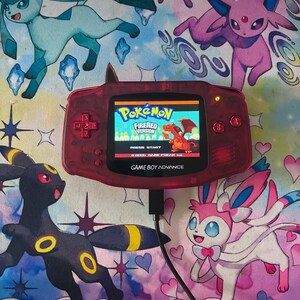 Op de afbeelding: Een doorschijnende rode Game Boy Advance handheld spelcomputer die het titelscherm van Pokémon FireRed Version met een Charizard-afbeelding weergeeft. Het apparaat heeft een D-pad, A- en B-knoppen en een zwarte oplaadkabel aangesloten.