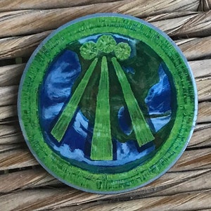 Könnte beinhalten: Eine runde, grüne und blaue Anstecknadel mit einem stilisierten Bild von drei grünen Stielen, die von einem Cluster aus drei grünen Kreisen ausstrahlen.