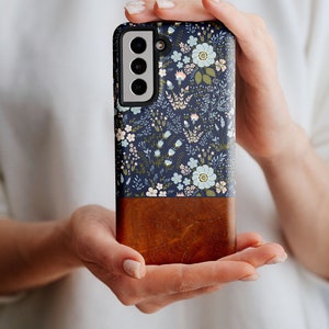 May include: A navy blue phone case with a floral pattern and a brown wood-grain bottom. The case has a raised camera bump with three lenses.
