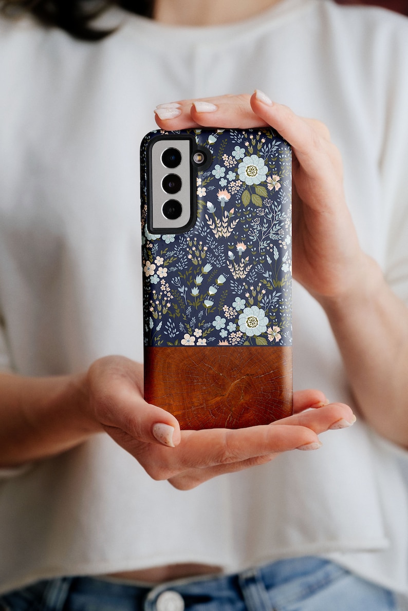 May include: A navy blue phone case with a floral pattern and a brown wood-grain bottom. The case has a raised camera bump with three lenses.