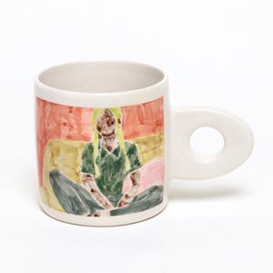 Puede incluir: Una taza de cerámica blanca con una ilustración pintada a mano de una figura con cabello largo y rubio sentada con las piernas cruzadas en un sofá amarillo sobre un fondo coral. El asa tiene una gran abertura circular en el centro.