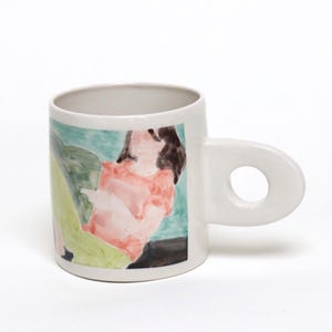 Puede incluir: Una taza de cerámica blanca con un asa grande y circular con un orificio central. La superficie exterior muestra una ilustración pintada en tonos verdes, rosas y negros, que representa una figura abstracta sobre un fondo claro.