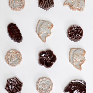 Puede incluir: Doce azulejos de cerámica con varios diseños, incluyendo flores, peces y patrones geométricos. Los azulejos son en tonos de marrón y blanco.