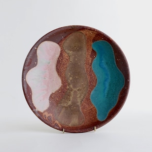 May include: A round ceramic plate with a speckled reddish-brown base glaze. The surface features three distinct, elongated, organic-shaped vertical patches of glaze in soft pink, muted brown, and vibrant teal, creating a layered, textured appearance.