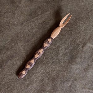 Puede incluir: Tenedor de madera con dos puntas. El mango es de color marrón claro con secciones estampadas en marrón oscuro. El tenedor probablemente se usa para servir o comer. El fondo es de color verde oscuro sólido.
