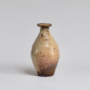 May include: A small, hand-thrown ceramic vase with a light brown glaze and a speckled finish. The vase has a rounded body and a narrow neck.