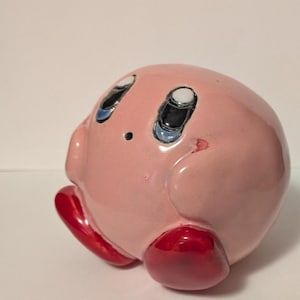 May include: A round, pink ceramic figurine of a character with large blue and black eyes, a small mouth, and two stubby arms. The figure has two shiny, bright red feet positioned at the bottom, giving it a rounded, spherical appearance.