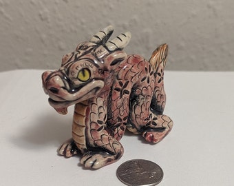 May include: A small, ceramic dragon figurine with a pink and brown patterned body. The dragon has a yellow eye and a white horn on its head.