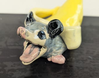 Könnte beinhalten: Eine Keramikfigur eines Opossums mit einem gelben Bananenschalenhut. Das Opossum ist grau und weiß mit schwarzen Augen und einer herausgestreckten rosa Zunge.
