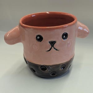 May include: A handmade ceramic mug with a pink face and dark brown bottom, resembling a cartoon character. The mug has two small ear-like handles and black eyes. The bottom has several dark brown circles.