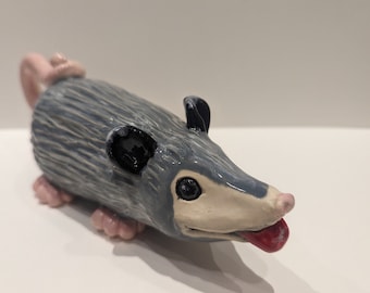 Könnte beinhalten: Eine kleine, graue Keramikfigur eines Opossums mit schwarzen Augen, einer rosa Nase und einer roten Zunge, die herausstreckt. Das Opossum hat einen weißen Bauch und einen strukturierten grauen Rücken.