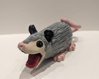 Könnte beinhalten: Eine kleine, graue und weiße Keramikfigur eines Opossums mit schwarzen Augen und einer herausgestreckten rosa Zunge. Die Figur hat eine strukturierte Oberfläche und rosa Füße.
