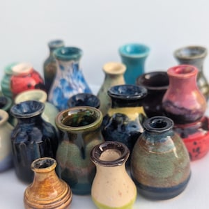 May include: A collection of small, handcrafted ceramic vases in various colors and designs. The vases feature a range of glazes, including blue, green, red, and brown, with unique patterns and textures. The vases are of varying heights and shapes.