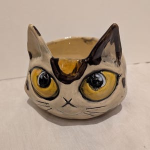 May include: A ceramic cat-shaped bowl with a cream-colored base and brown and black accents. The cat's face features large yellow eyes, a black nose, and whiskers. The ears are pointed and the bowl is open at the top.