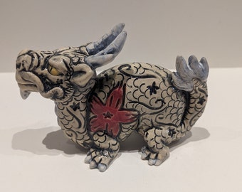 May include: A small ceramic dragon figurine with a white glaze and black and red painted details. The dragon has a stylized design with scales and a flower on its side.