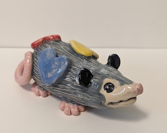 Könnte beinhalten: Eine handbemalte Keramikfigur eines Opossums mit grauem Körper, rosa Ohren und einem rosa Schwanz. Das Opossum hat ein blaues Herz auf dem Rücken, gelbe und rote Akzente und schwarze Augen.
