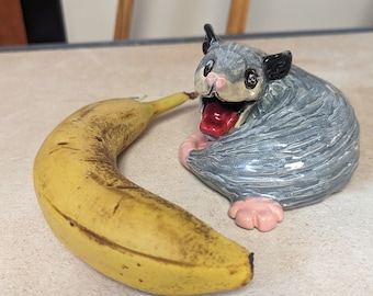 Könnte beinhalten: Eine Keramikfigur eines grauen Zuckergleiters mit herausgestreckter roter Zunge, die neben einer gelben Banane auf einer weißen Oberfläche sitzt.