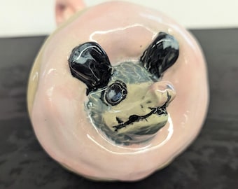 Könnte beinhalten: Eine Keramikfigur eines Opossums mit schwarzen Ohren und einer rosa Glasur. Das Gesicht des Opossums ist mit schwarzen Augen und einer kleinen Nase detailliert.