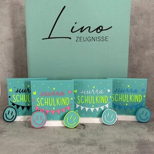 Könnte beinhalten: Ein großer mintgrüner Ordner mit der Aufschrift Lino ZEUGNISSE, dazu vier kleine quadratische mintgrüne Schilder mit dem Text Hurra SCHULKIND, bunten Wimpelketten und Sternen sowie fünf runde, farbenfrohe Smiley-Schlüsselanhänger.