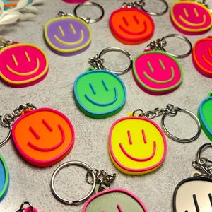 Könnte beinhalten: Eine Sammlung runder Schlüsselanhänger mit einem erhabenen Smiley-Gesicht. Jeder Schlüsselanhänger besteht aus einer leuchtend farbigen Basis mit kontrastierendem Rand und einem passenden Smiley-Motiv, befestigt an einem silberfarbenen Metallring.