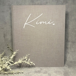 Könnte beinhalten: Ein hellgraues, strukturiertes Buch mit dem Namen "Kimi" in weißer Schrift. Ein kleines weißes Herz folgt dem Namen. Das Buch ist vor einem grauen Hintergrund mit einem Zweig weißer Blätter in der unteren linken Ecke platziert.