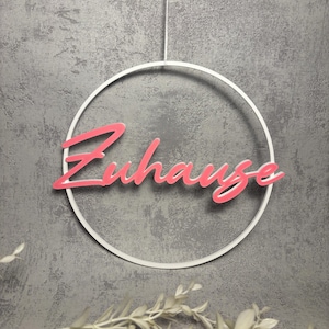 Könnte beinhalten: Ein weißer, kreisförmiger Wandbehang mit dem Wort "Zuhause" in rosa Schrift. Das Wort ist innerhalb des Kreises zentriert, der an einer dünnen weißen Schnur vor einem grau strukturierten Hintergrund hängt.