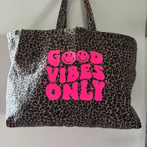Könnte beinhalten: Eine Tragetasche mit braun-schwarzem Leopardenmuster. Auf der Vorderseite befindet sich der Schriftzug "GOOD VIBES ONLY" in leuchtendem Pink, wobei ein Smiley in den Buchstaben O integriert ist und kleine Sterne das Textdesign ergänzen.