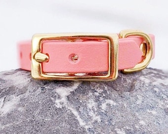 May include: A pink leather dog collar with a gold buckle. The collar is on a gray rock.
