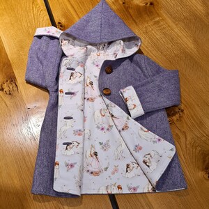 May include: Purple hooded jacket with a white lining featuring a unicorn and floral print. The jacket has a button closure and a herringbone pattern.