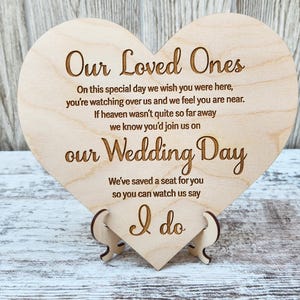 May include: A wooden heart-shaped sign with the text "Our Loved Ones" and a message about remembering loved ones on a wedding day. The sign is on a stand and reads "On this special day we wish you were here, you're watching over us and we feel you are near. If heaven wasn't quite so far away we know you'd join us on our Wedding Day. We've saved a seat for you so you can watch us say I do."