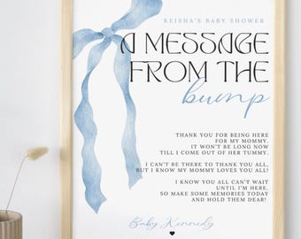 May include: A framed baby shower sign with a light wood frame. The sign features a blue ribbon design and text that reads "A Message From The Bump." The text includes a message from the baby, the baby's name, and the event details.