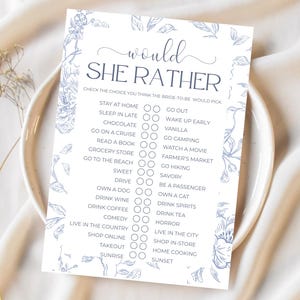 Peut inclure: Une carte blanche ornée de motifs floraux bleus et du texte "Would She Rather". La carte propose un jeu pour une fête prénuptiale, avec des choix comme "Rester à la maison" et "Sortir".