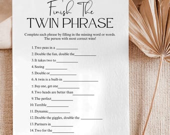 May include: A white card with the text "Finish The TWIN PHRASE" in elegant script. The card features fill-in-the-blank phrases related to twins, designed as a game. The background is a neutral beige, with a dried palm leaf.