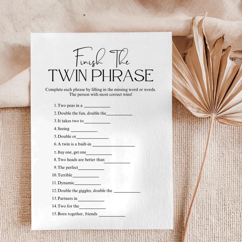 A white baby shower game card with the text "Finish The TWIN PHRASE" in elegant script. The card features fill-in-the-blank phrases related to twins, designed as a game. The background is a neutral beige, with a dried palm leaf.