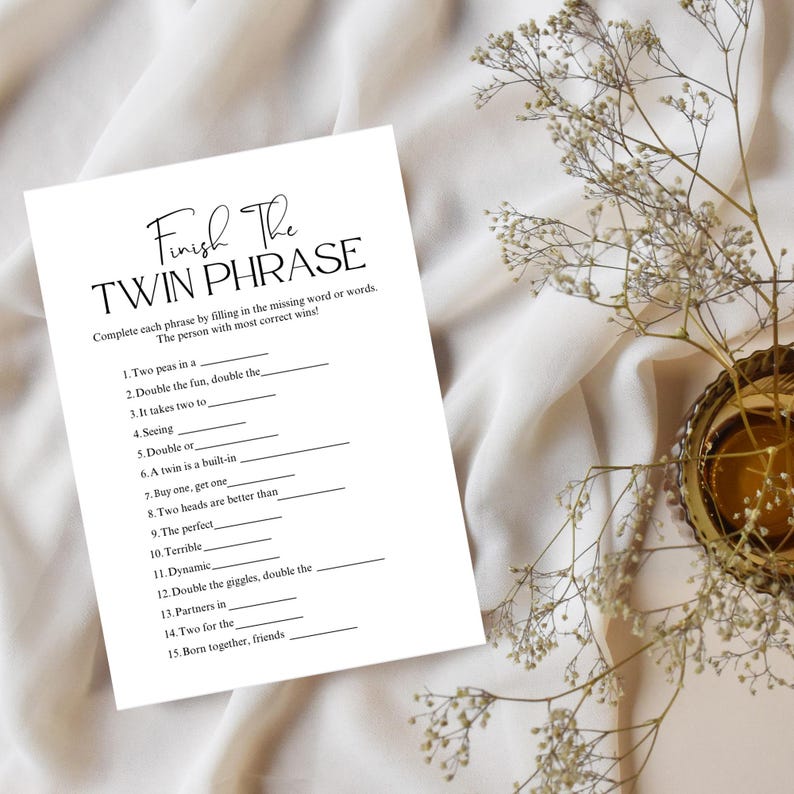 May include: A white paper with the text "Finish The Twin Phrase" in elegant script. The game sheet includes fill-in-the-blank phrases. The paper is on a white fabric surface, with floral and gold accents.