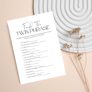 May include: A white paper game sheet titled "Finish The Twin Phrase". The game challenges players to complete phrases. The sheet is on a neutral background with decorative elements, suitable for a bridal shower or baby shower activity.
